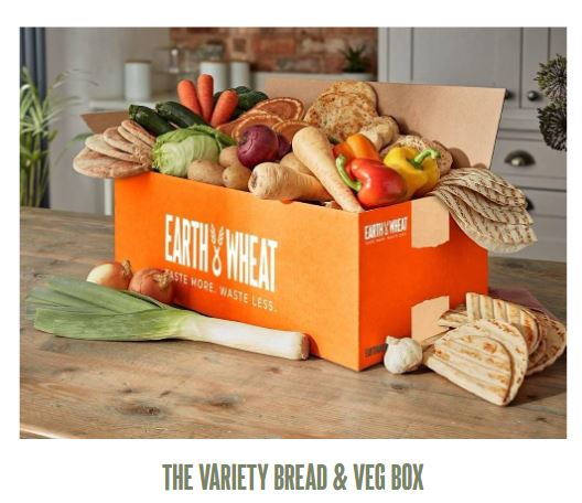 An orange box labeled "Earth & Wheat" filled with assorted breads and vegetables, including peppers, leeks, and various loaves, sits on a wooden table.