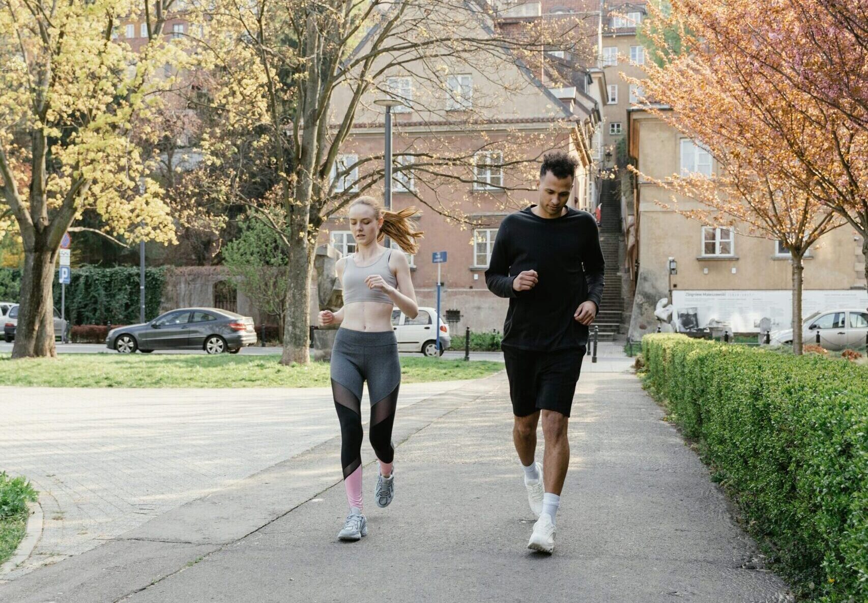 Two people jogging on a paved path in a park, surrounded by trees and buildings in the background.