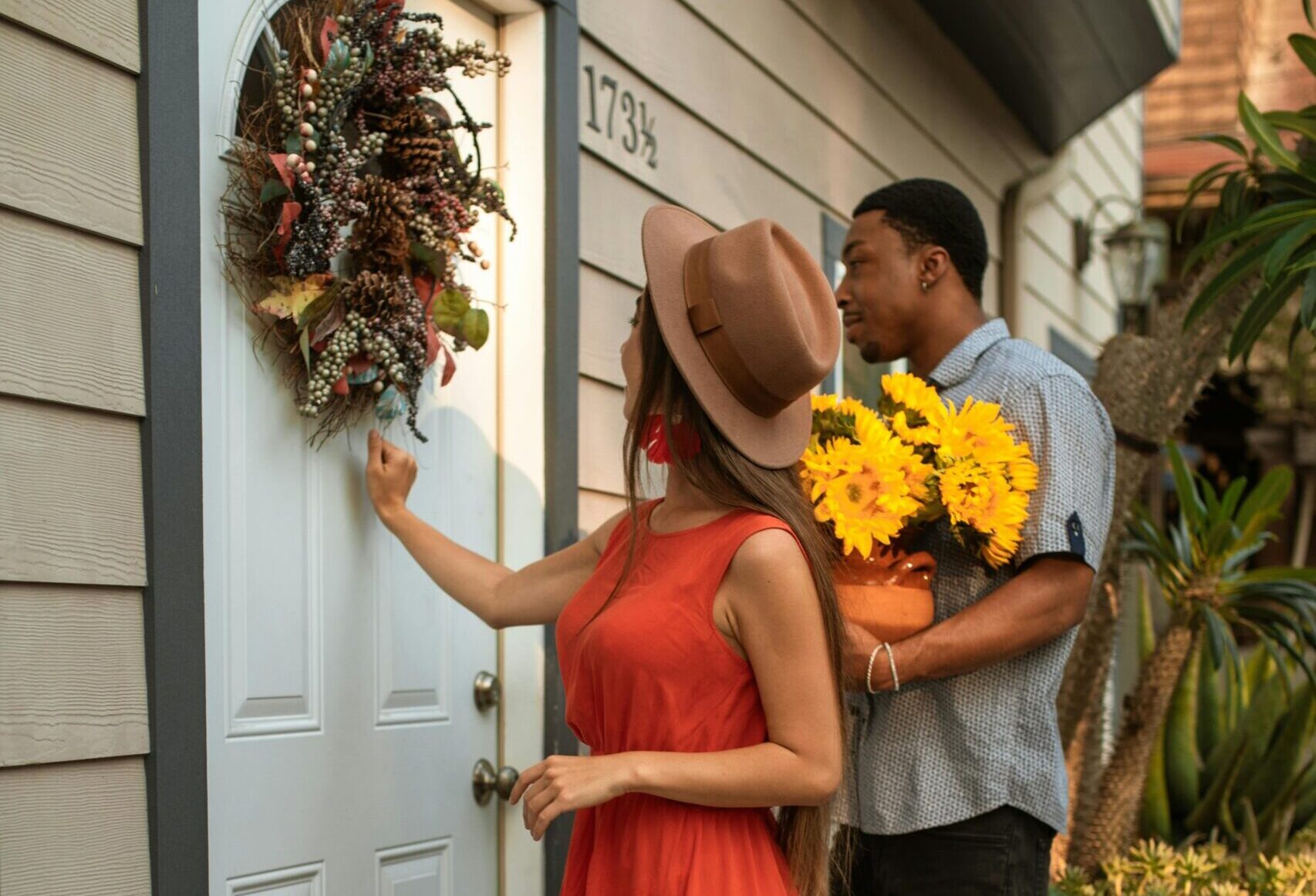 A woman in a red dress knocks on a front door, while a man beside her holds a bouquet of sunflowers.