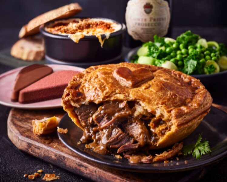 A meat pie on a plate with a cut piece, surrounded by broccoli, mashed potatoes, a dish with melted cheese, and a bottle of sparkling wine.