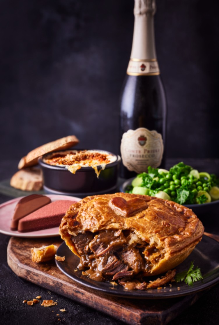 A meat pie on a plate, with a bottle of champagne, a dish of vegetables, a small casserole, and decorative plates in the background.