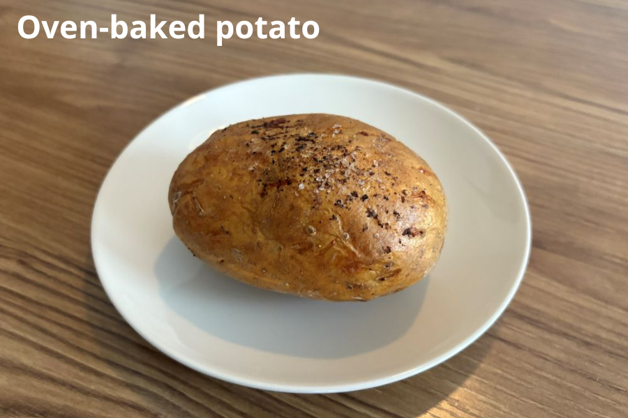 A whole oven-baked potato on a white plate, seasoned with black pepper, on a wooden surface.