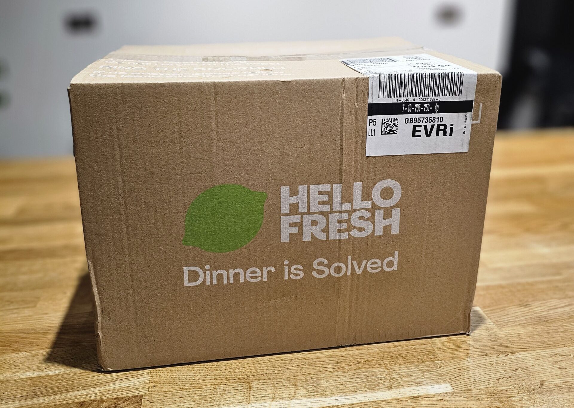 A HelloFresh delivery box sits on a wooden table. The box displays the logo and the phrase "Dinner is Solved.