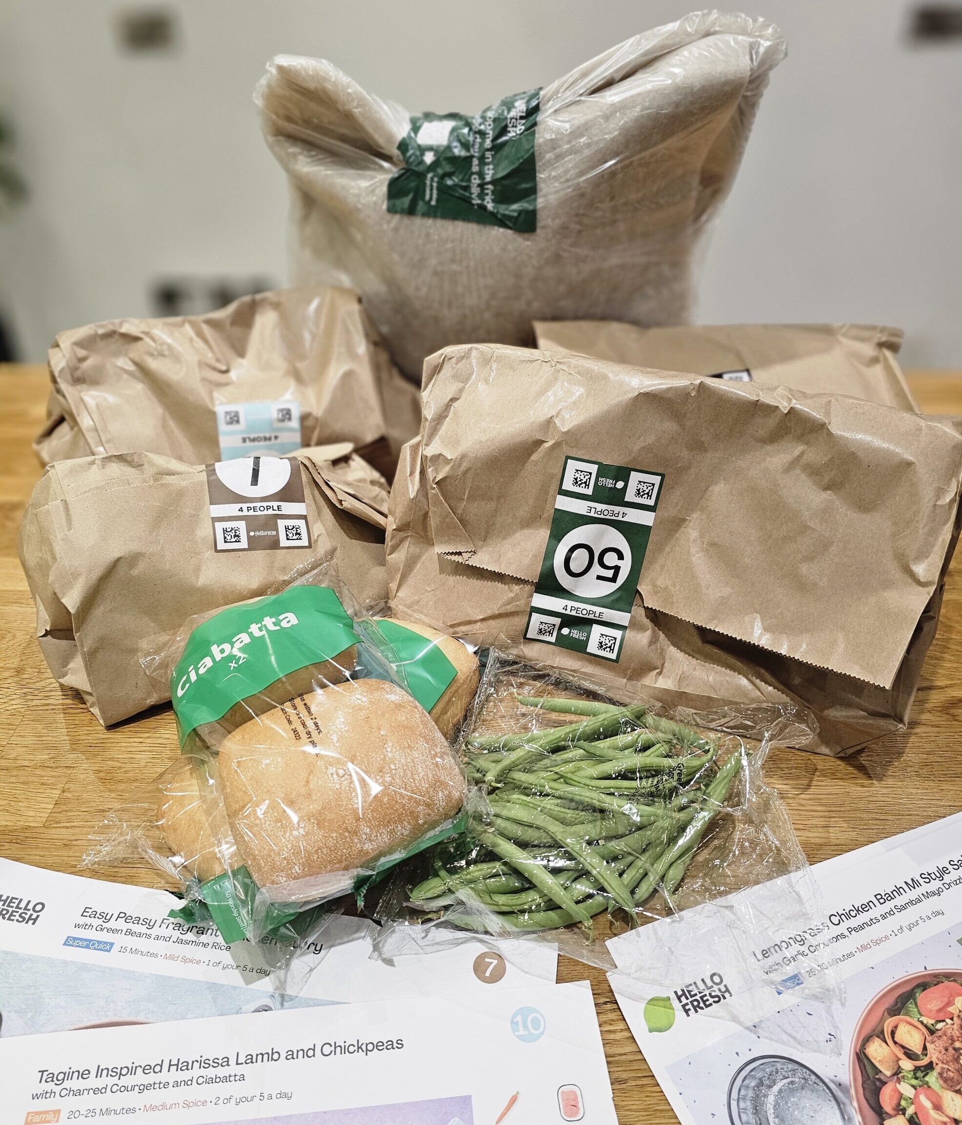 Several meal kit packages, green beans, ciabatta rolls, and recipe cards are arranged on a wooden surface.