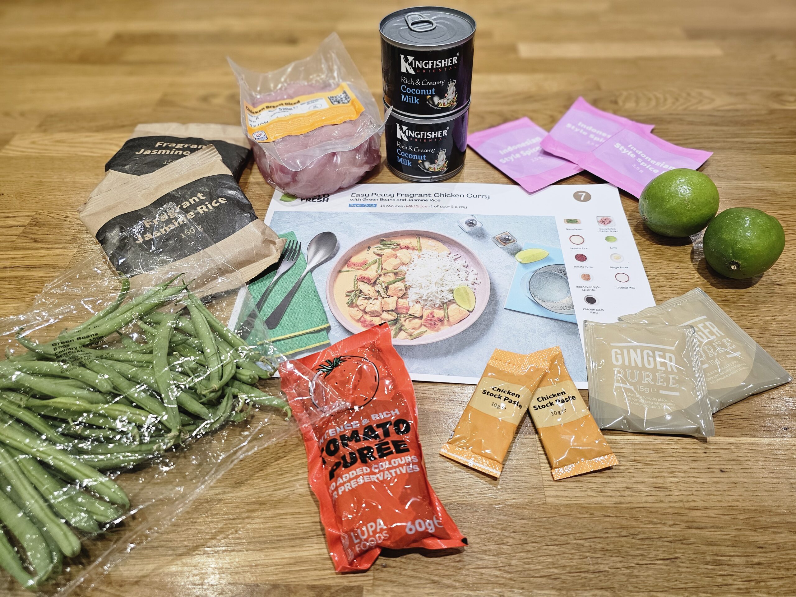 A selection of ingredients for making chicken curry, including chicken, coconut milk, ginger paste, green beans, rice, curry paste, limes, and recipe card, arranged on a wooden surface.