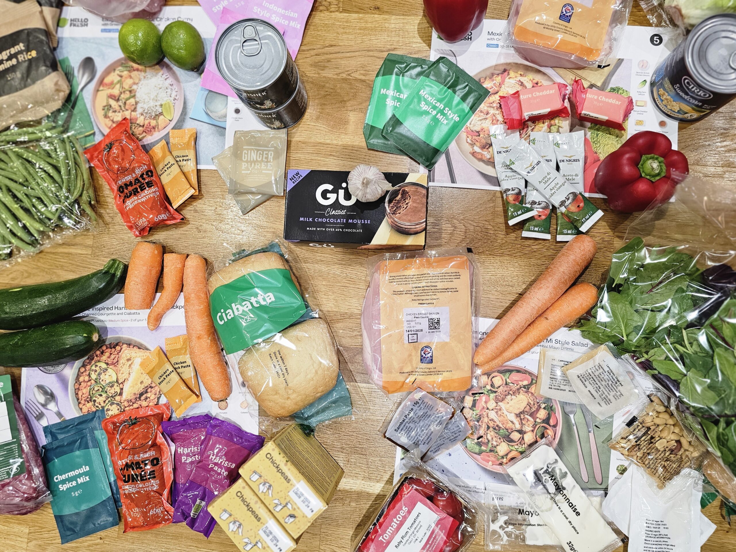 A variety of groceries are spread out on a wooden table, including vegetables, canned goods, packaged snacks, and bread. Recipe cards are also visible.