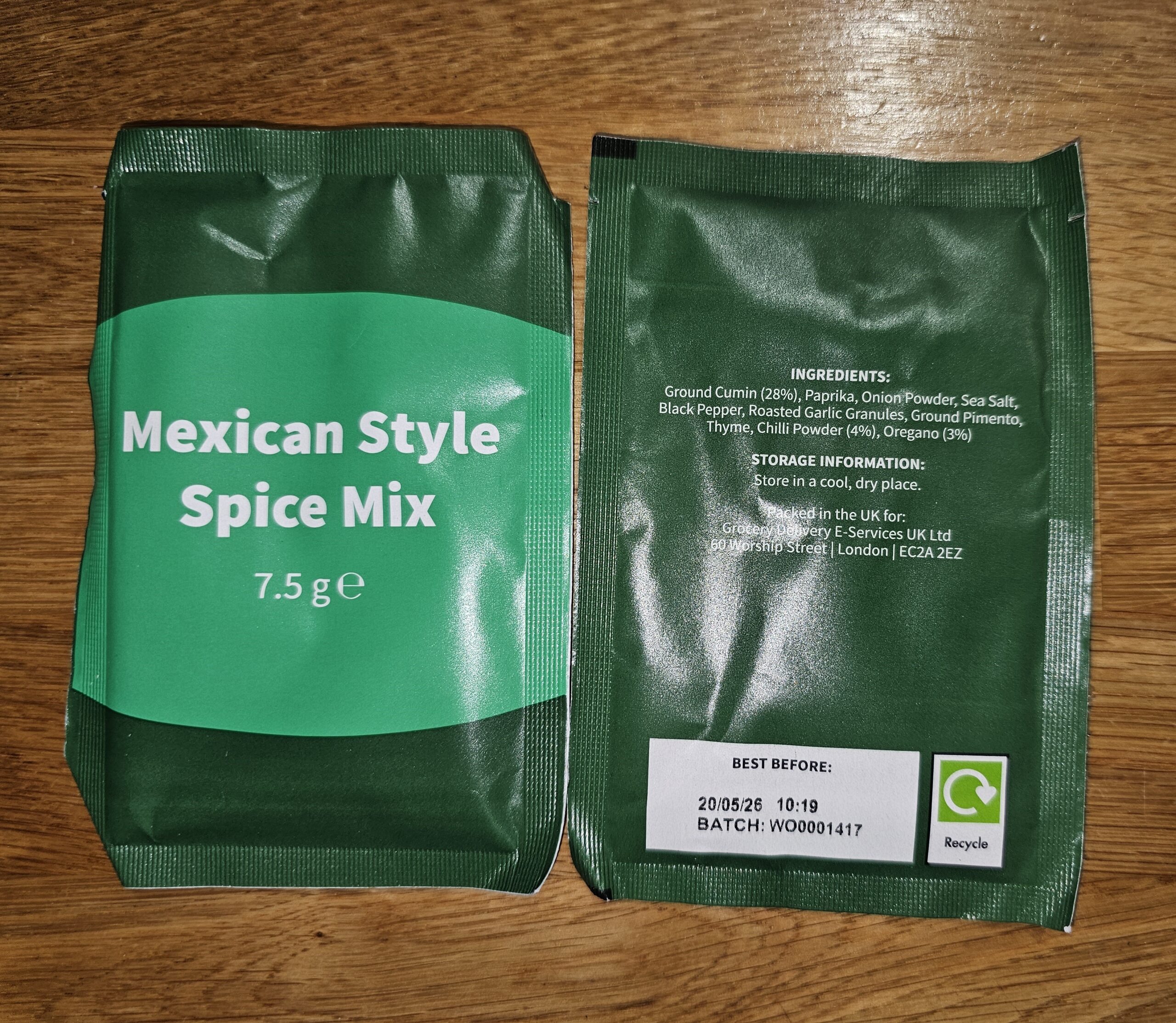 Front and back of a green spice mix packet displaying product name, weight, ingredients, and expiration date on a wooden surface.