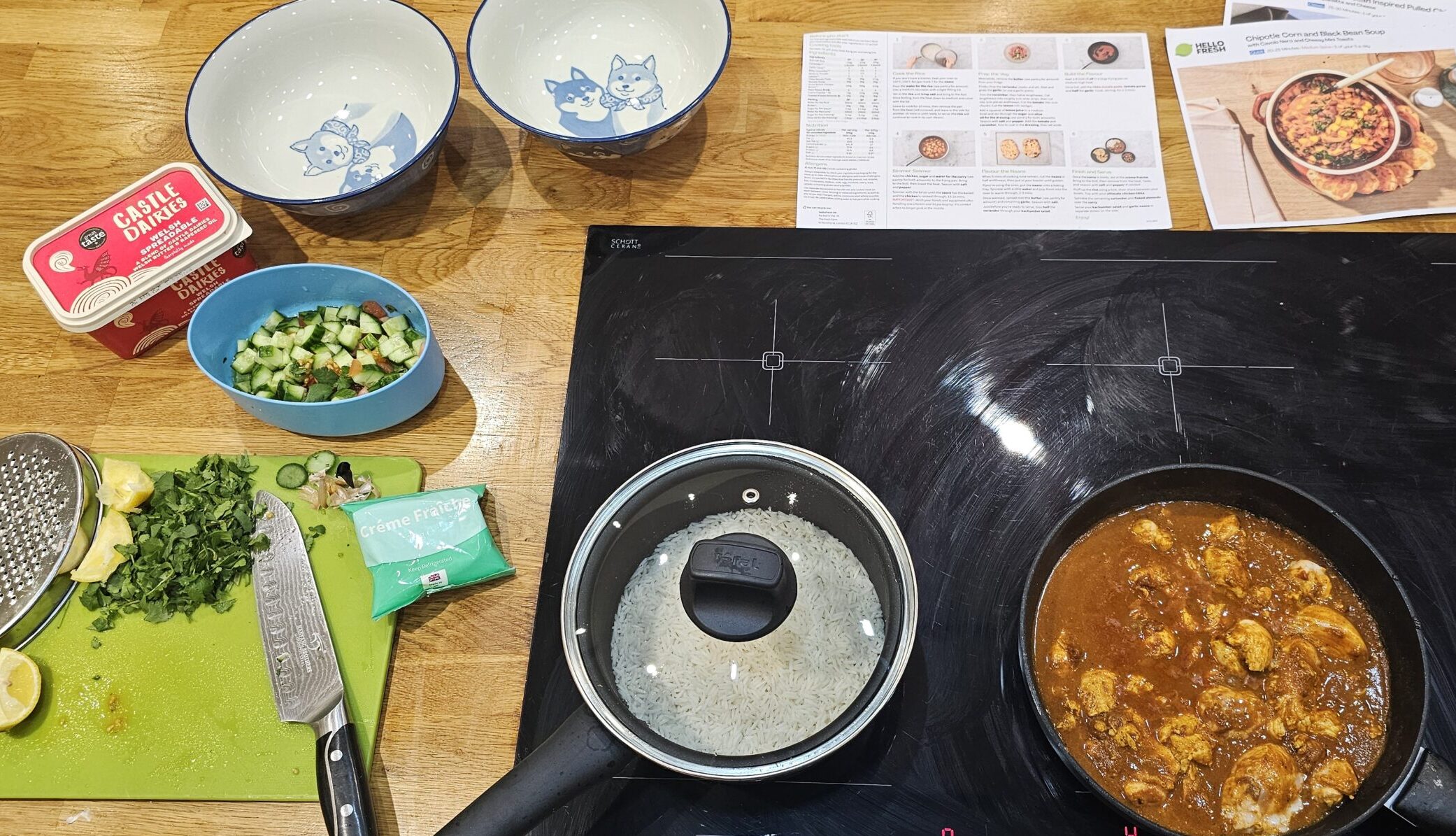 A stovetop with a pan of curry next to a pot with a lid, surrounded by chopped herbs, a knife, seasoning, two empty bowls, and recipe cards on a wooden counter.