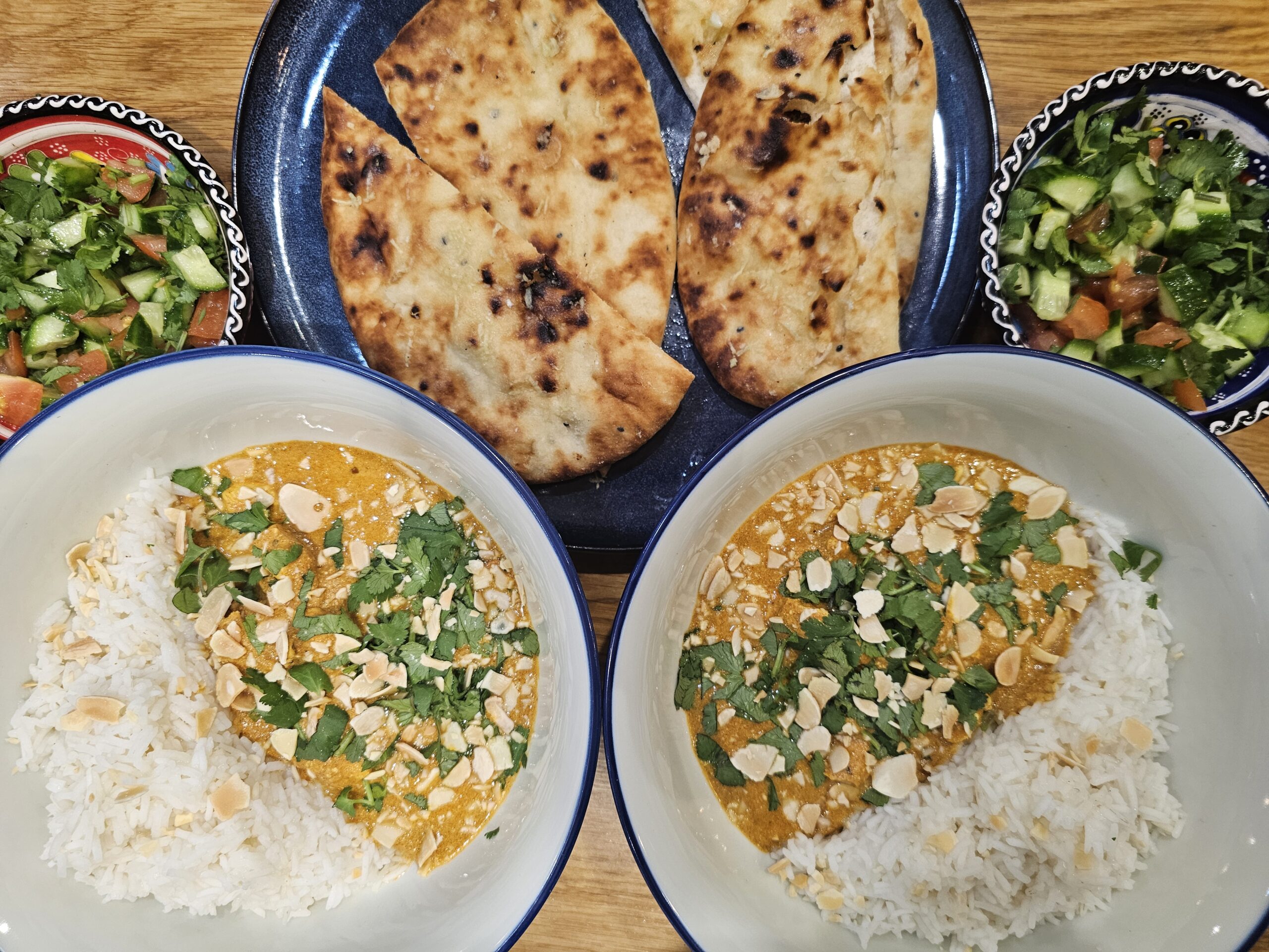 Two bowls of curry with rice topped with cilantro and almonds, two naan breads, and two bowls of chopped salad with cucumber and tomatoes on a wooden table.