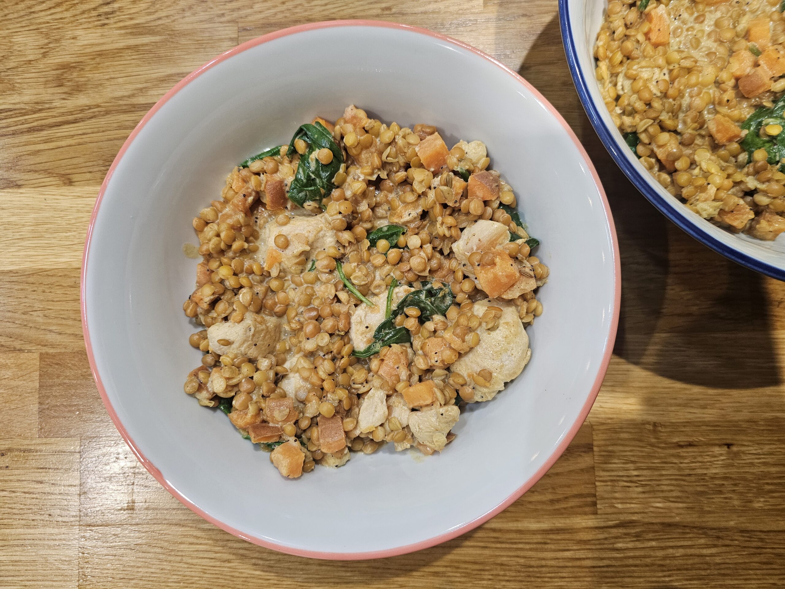 A bowl of lentil salad with spinach, sweet potato, and chicken pieces on a wooden surface. Another partially visible bowl is in the background.