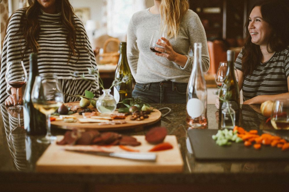 Four people stand around a kitchen island with wine and appetizers, including meat and vegetables, on wooden boards.