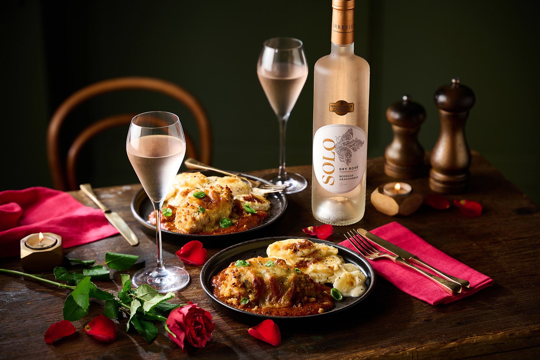 Two plates of lasagna, a bottle of white wine, wine glasses, red napkins, roses, candles, and salt and pepper shakers on a wooden table.