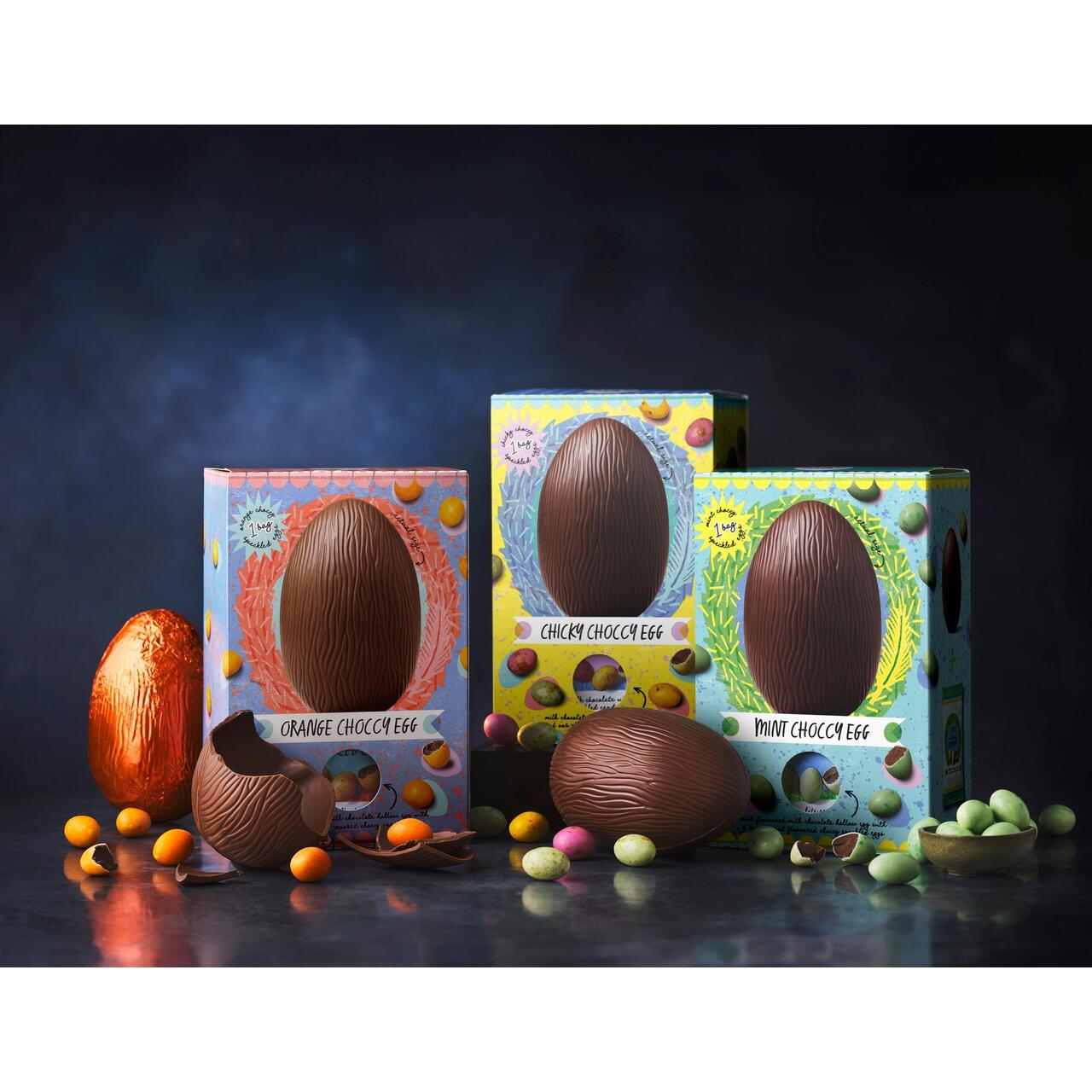 Three boxed chocolate eggs labeled "Orange Choccy Egg," "Choccy Choccy Egg," and "Mint Choccy Egg," surrounded by colorful candy and one unboxed egg.