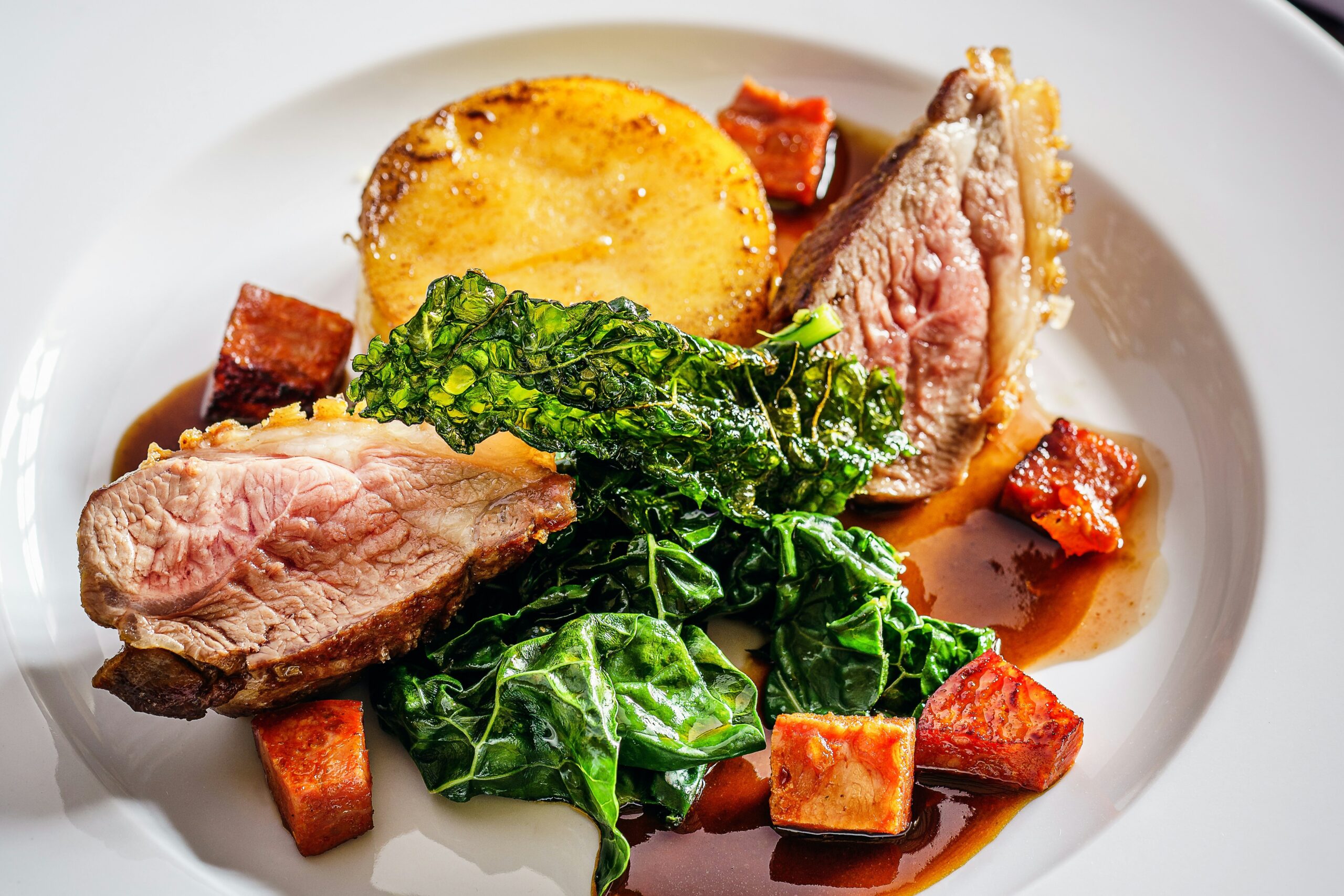 A plated dish featuring sliced duck, kale, a round potato, cubes of sweet potato, and sauce, arranged on a white plate.