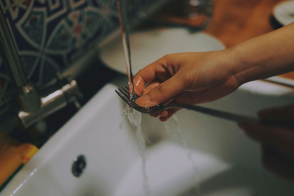 A person rinses a fork under running water in a kitchen sink.