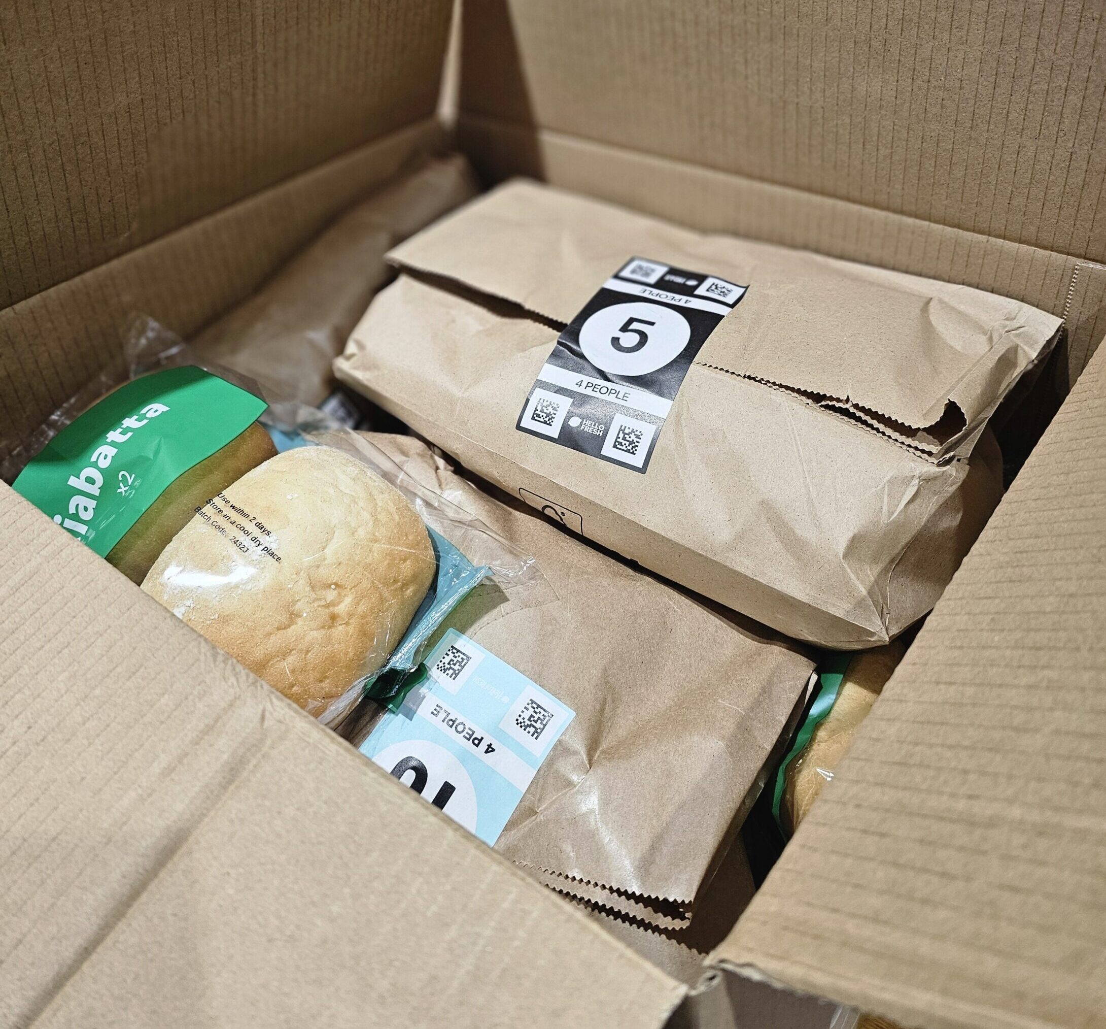 A cardboard box containing brown paper bags labeled with numbers, partially showing packaged ciabatta bread inside.