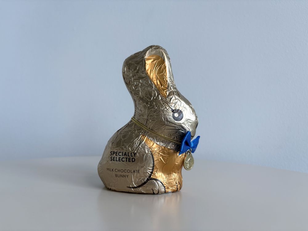 Gold-foil wrapped chocolate bunny labeled "Specially Selected Milk Chocolate Bunny" with a blue ribbon sitting on a white surface.