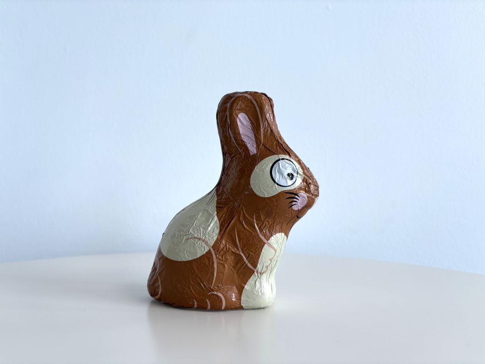 Chocolate rabbit wrapped in brown foil with white spots and cartoon-like eyes, sitting on a white surface against a light blue background.