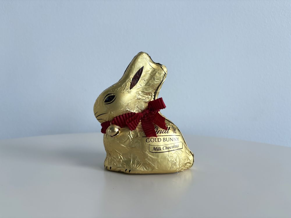 A gold-foil wrapped chocolate bunny with a red ribbon and bell sits on a white surface against a blue background.