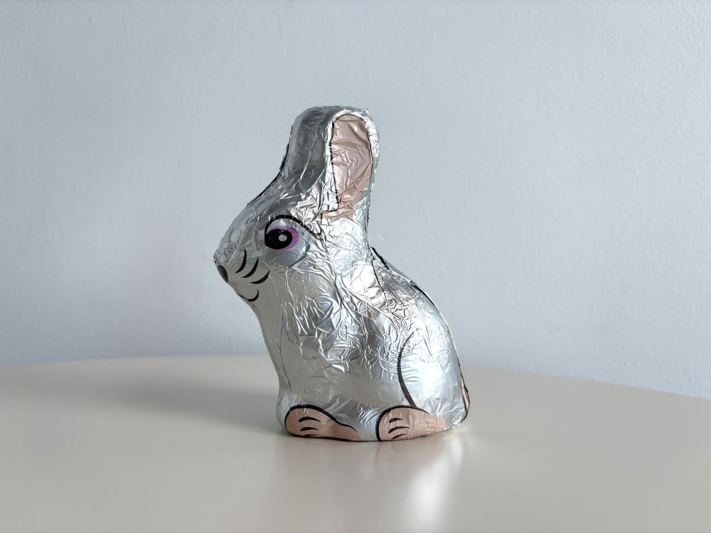 Silver foil-wrapped chocolate bunny with pink ears and an eye design, sitting on a round surface against a light blue background.