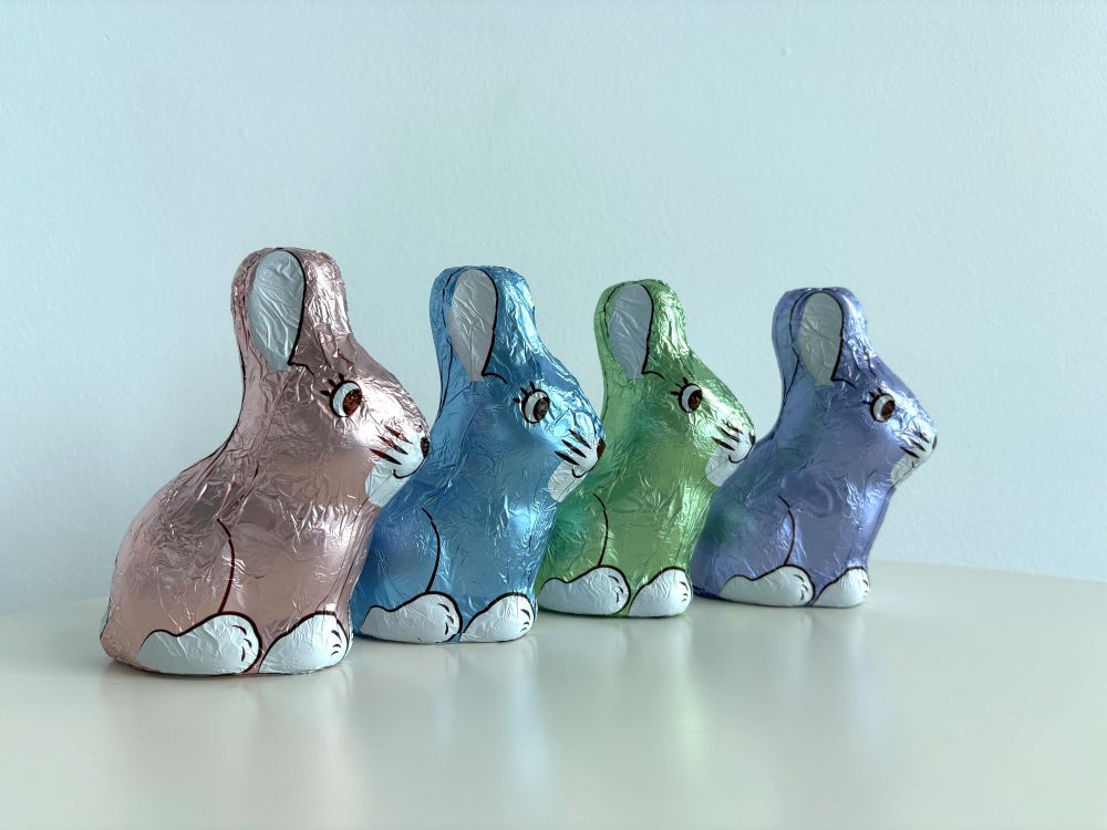 Four foil-wrapped chocolate bunnies in pink, blue, green, and purple lined up on a white surface against a pale blue background.